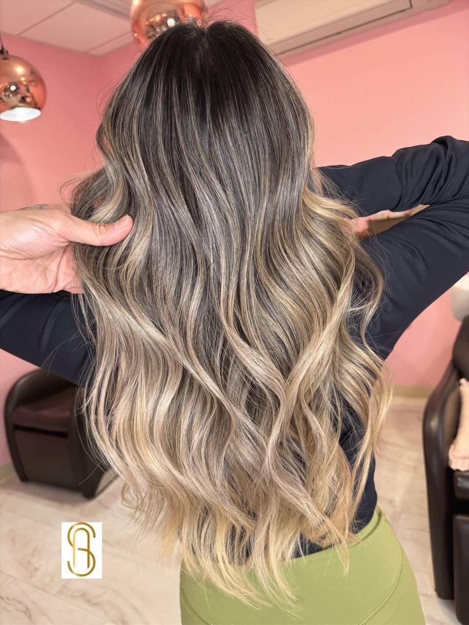 Long, wavy hair styled with a dark brown to blonde balayage ombre color, viewed from the back in a salon setting.