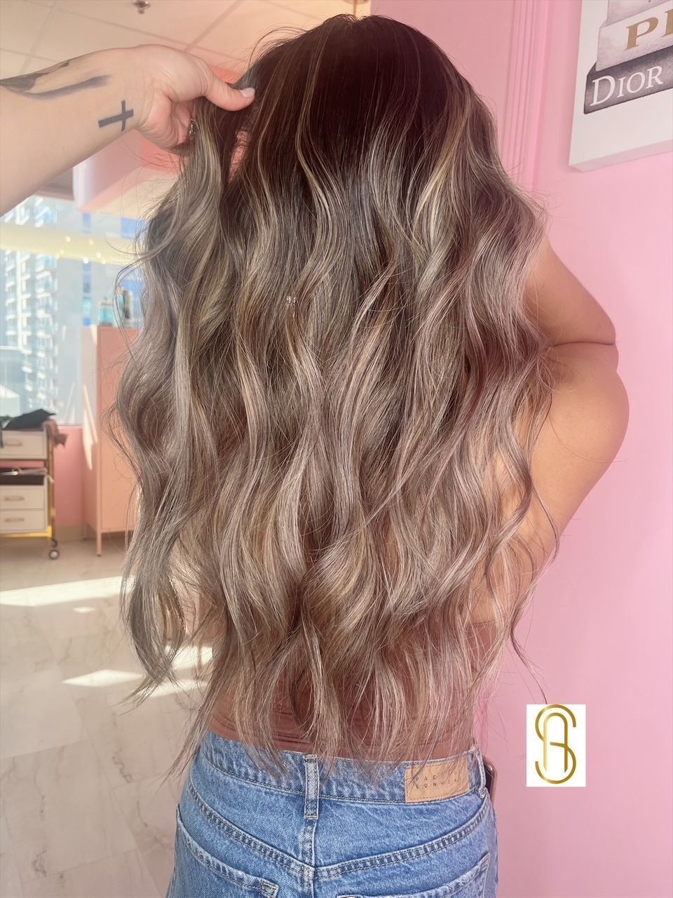 Long, wavy, brunette hair with highlights, viewed from behind in a room with pink walls and a gold salon logo.