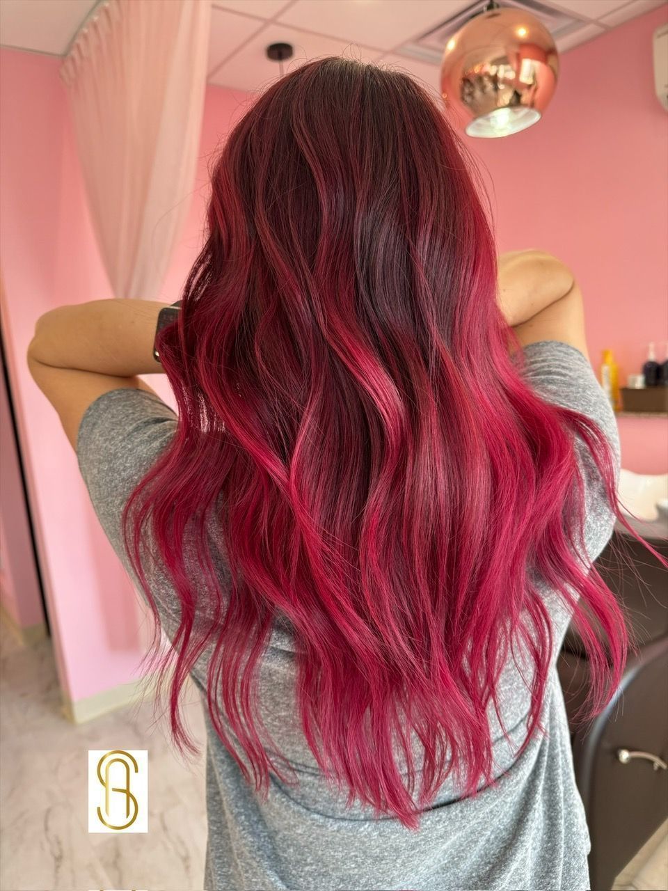 A person with long, wavy hair colored in vibrant magenta and deep red tones, seen from behind in a salon setting.