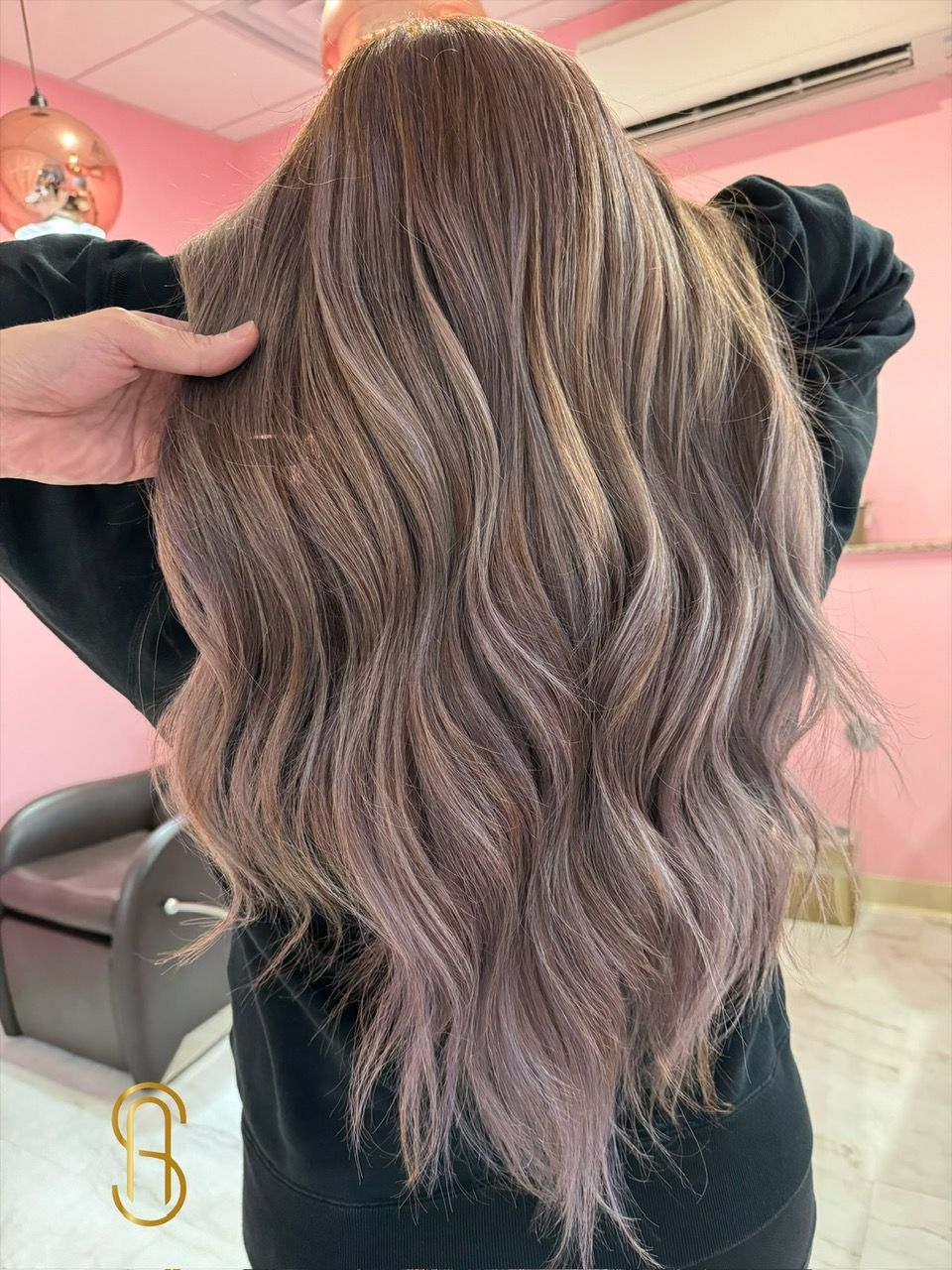 A person with wavy, multi-tonal brown and ash-blonde hair shown from behind in a salon setting.