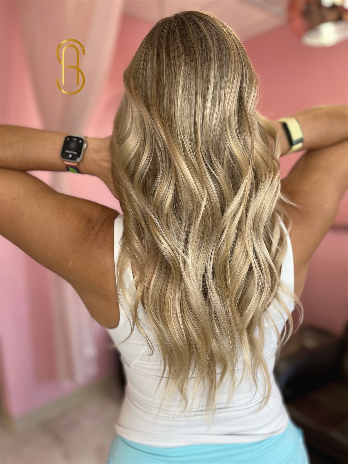 A person from behind shows off long, wavy, blonde hair with highlights, wearing a white tank top and two smartwatches.