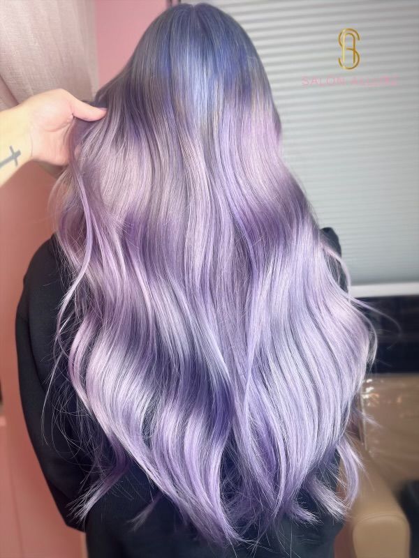Long, wavy hair colored in a blend of pastel purple and lavender shades, with a hand holding a section near the top.