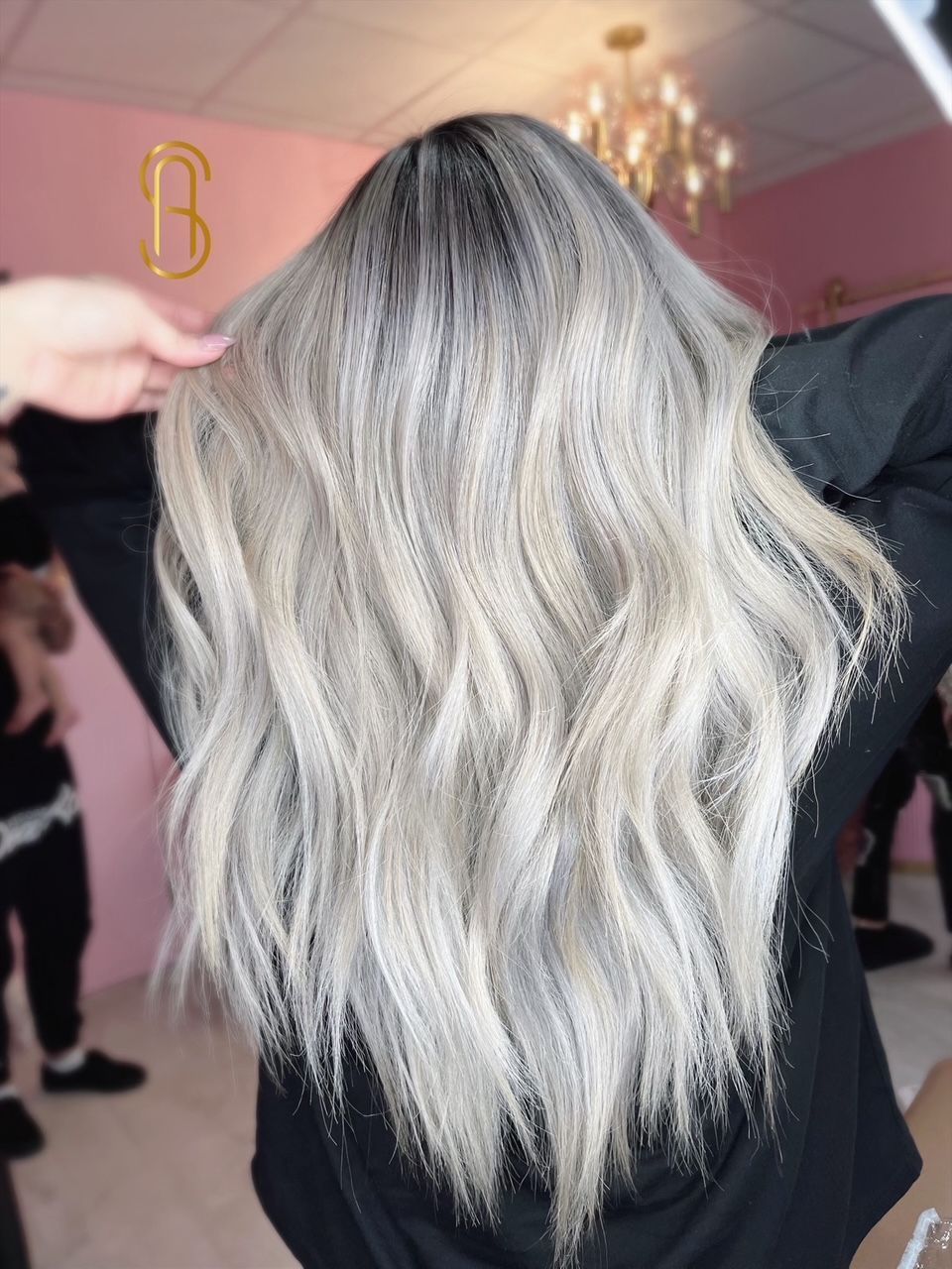 Long, wavy, ash-blonde hair with a dark root melt, viewed from the back in a salon setting.