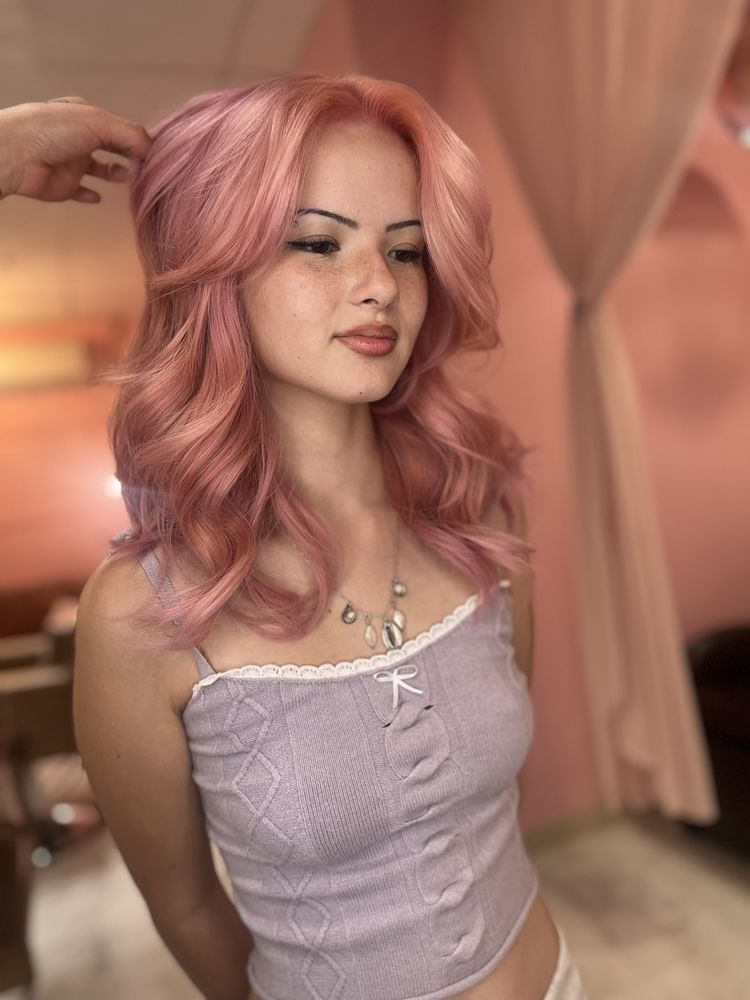 A person with wavy, pastel-pink hair and curtain bangs, wearing a lavender ribbed tank top and a silver necklace.