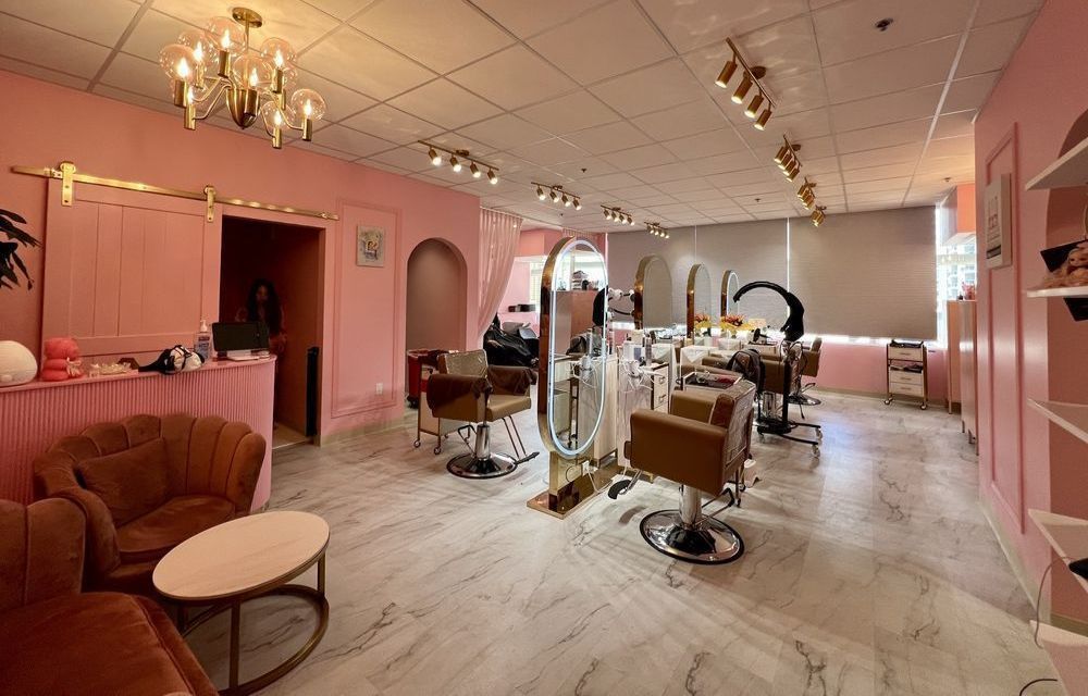 A modern hair salon with pink walls, marble floors, gold accents, a velvet couch, and multiple styling stations.
