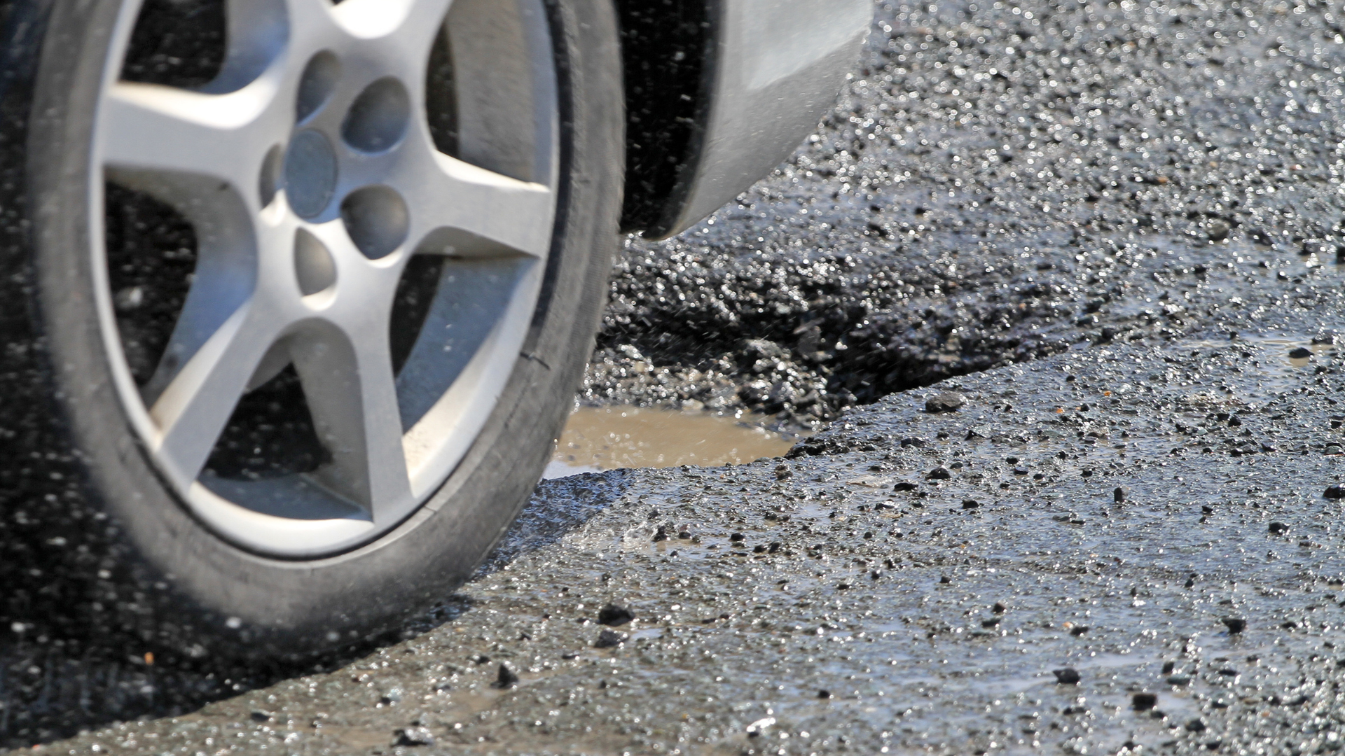 Car wheel beside a large pothole filled with muddy water on a damaged road
