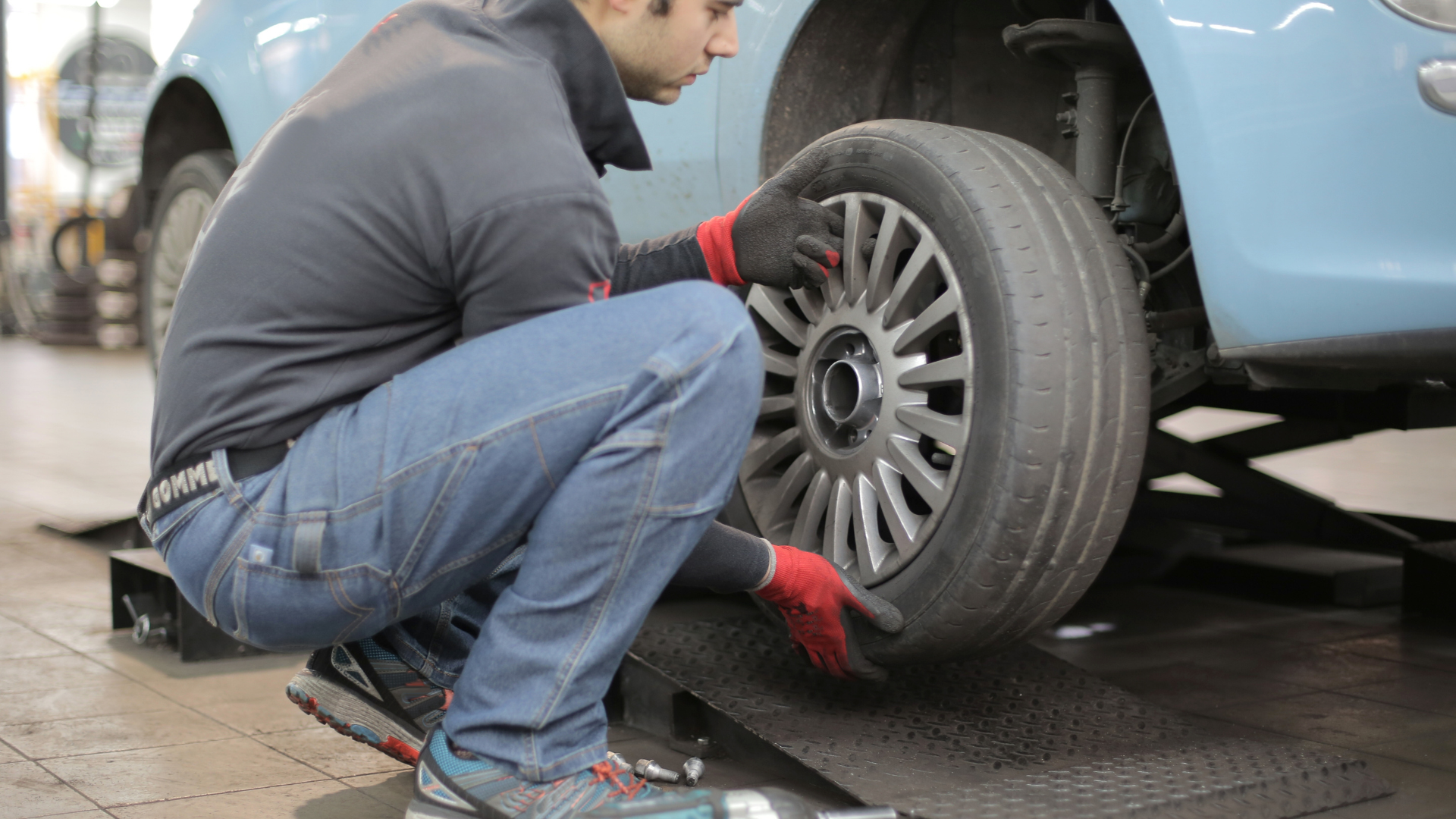 Mechanic kneeling beside a light blue car, checking or replacing a front wheel in a garage.