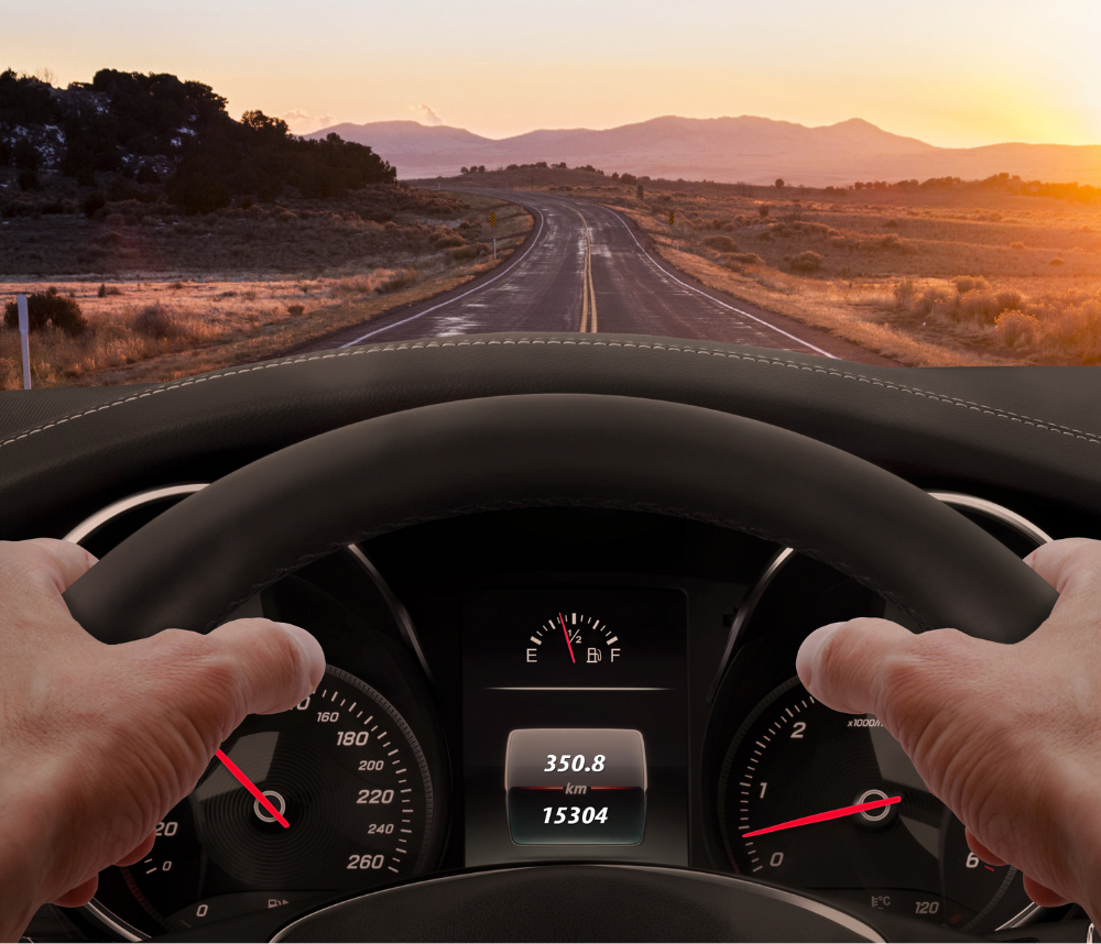 Hands gripping a steering wheel, driving on a road towards a sunset. Dashboard visible, with speed and mileage displayed.
