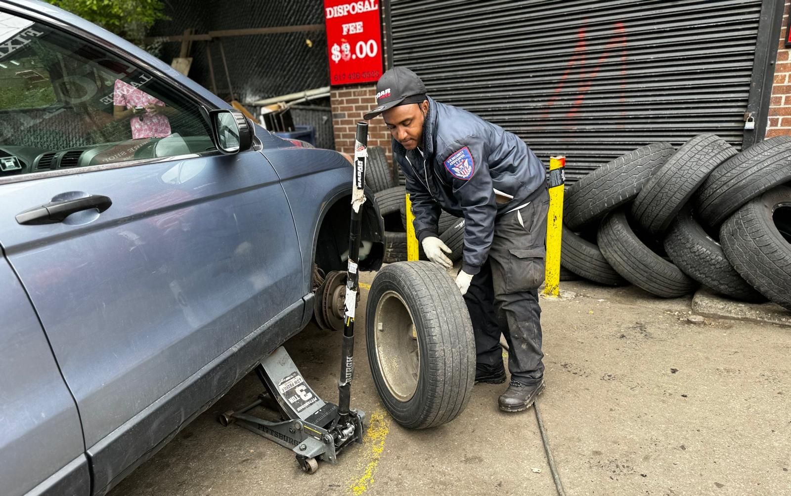 A man is changing a tire on a car in front of a pile of tires.