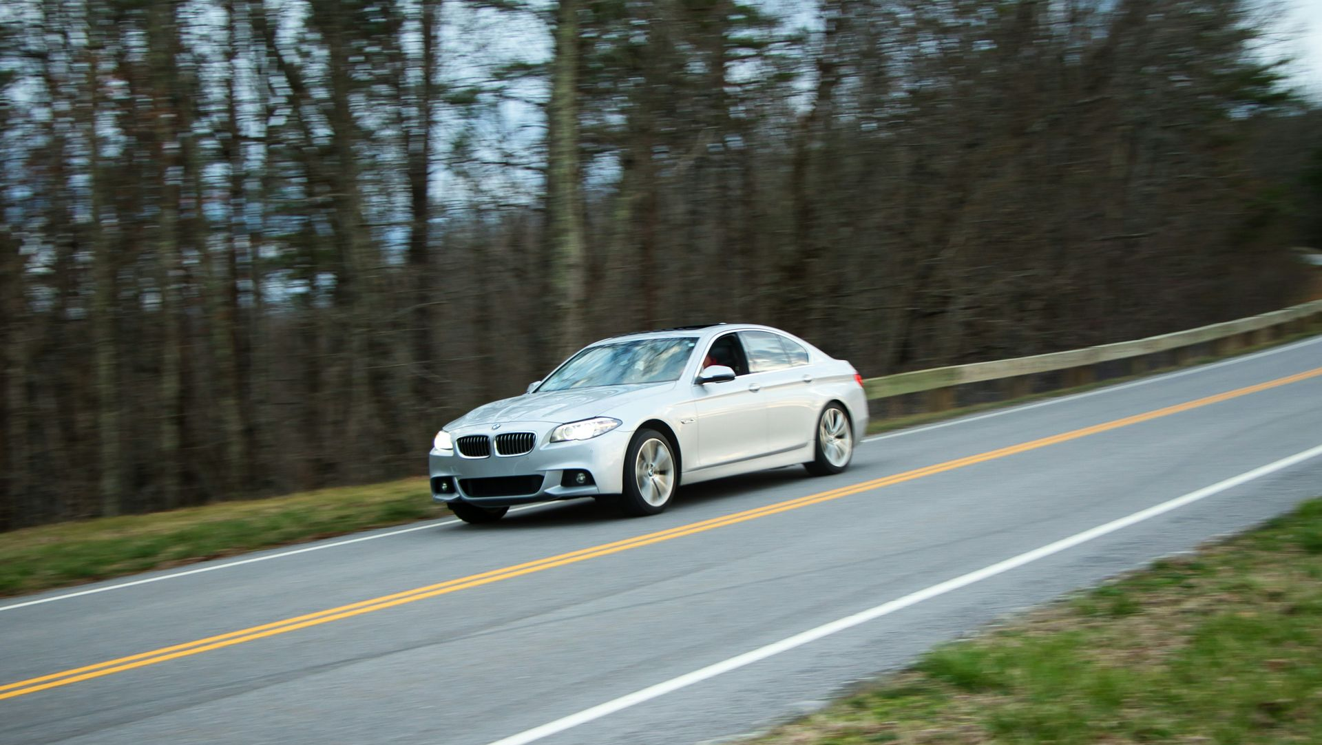 Silver sedan driving on a curving road beside a wooded area