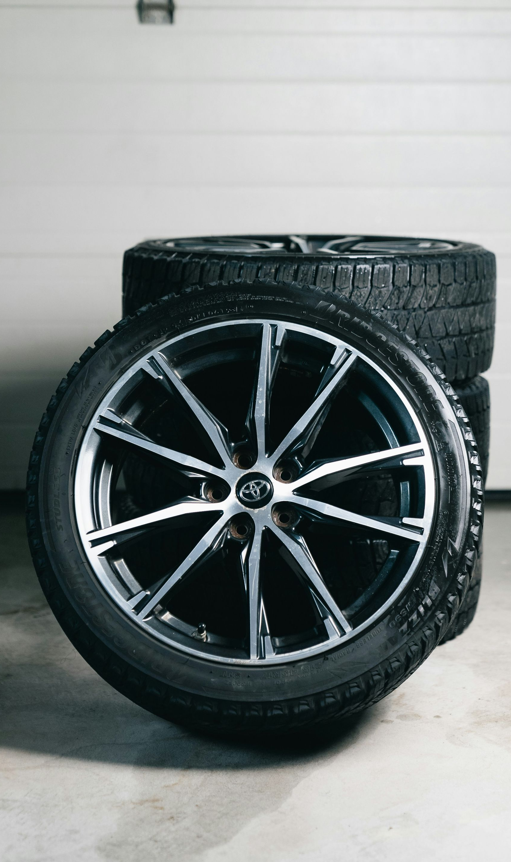 A stack of black tires with sleek, silver-accented alloy rims resting on a concrete floor in a garage.
