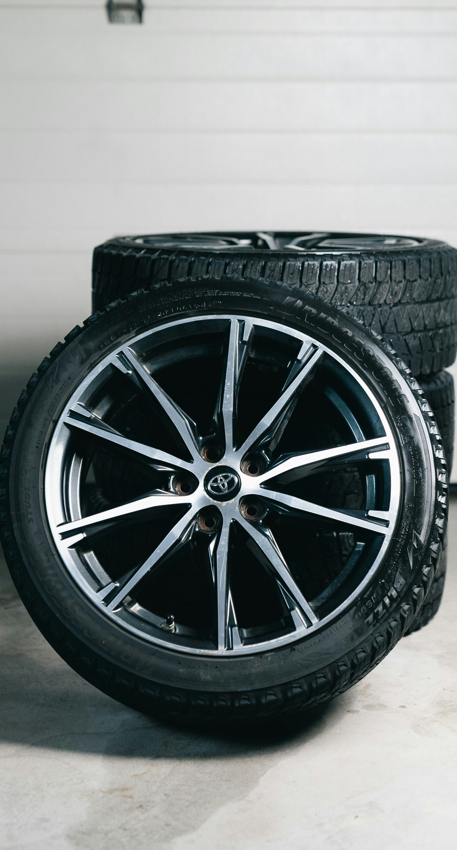 A stack of black tires with sleek, silver-accented alloy rims resting on a concrete floor in a garage.