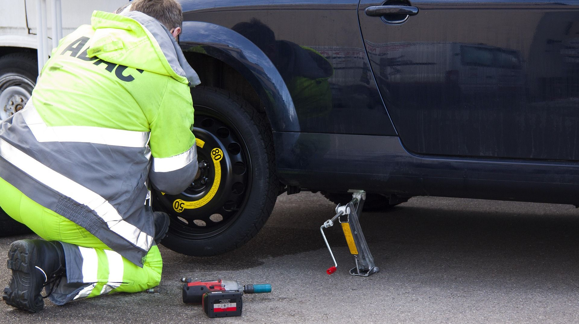 Roadside worker in yellow vest changing a tire on a dark blue car, using tools on pavement.