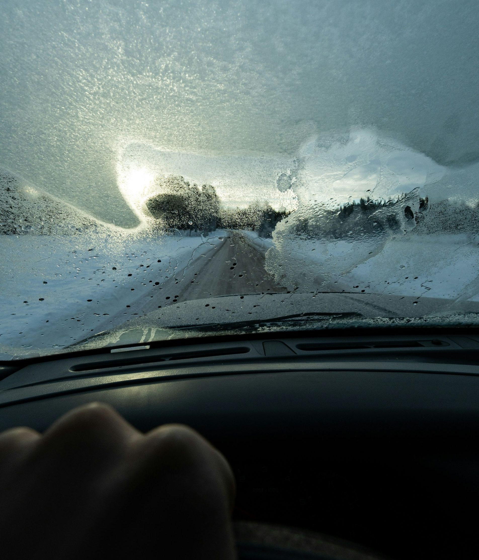 View from a car windshield covered in ice and frost, with a hand on the steering wheel, driving on a snowy road.