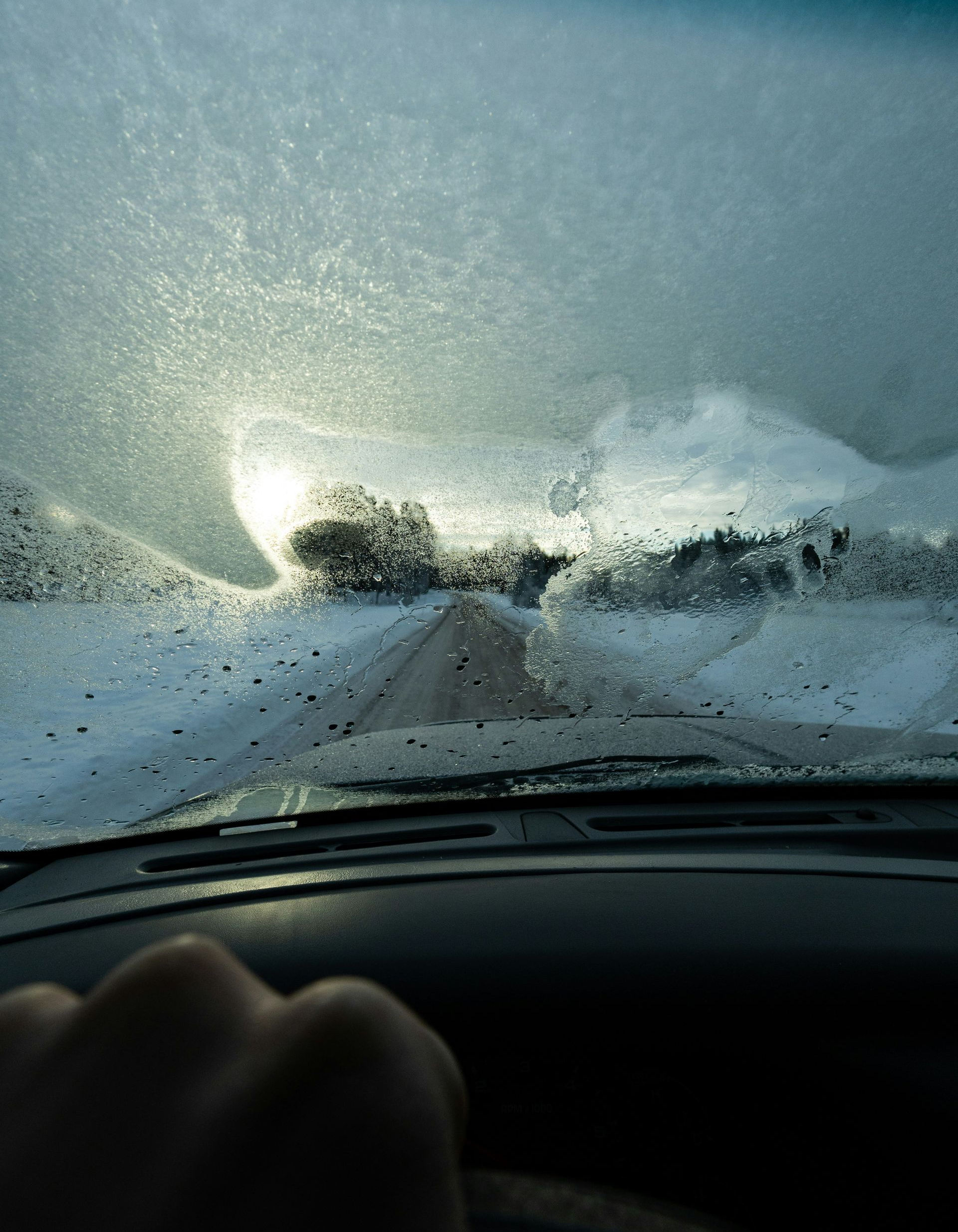 View from a car windshield covered in ice and frost, with a hand on the steering wheel, driving on a snowy road.