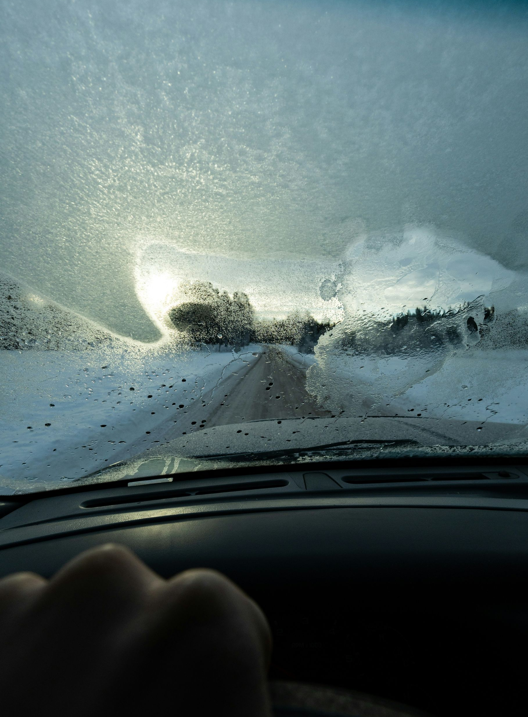 View from a car windshield covered in ice and frost, with a hand on the steering wheel, driving on a snowy road.