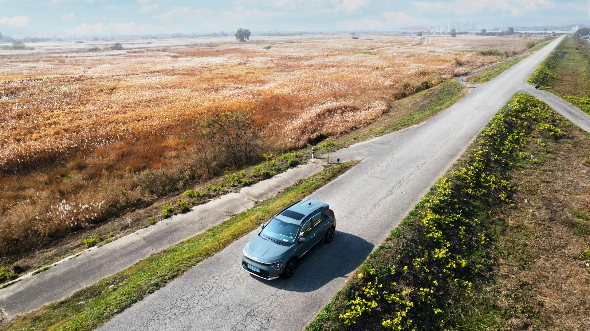 Dark green car driving on a narrow road beside a dry open field under a clear sky