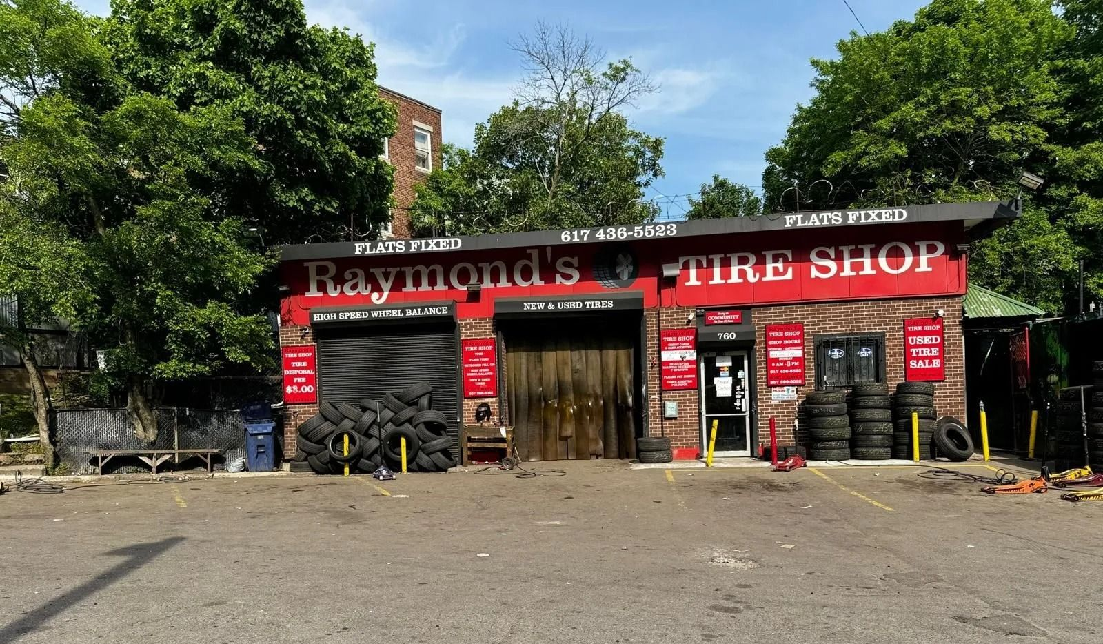 Raymond's Tire Shop exterior with tires stacked. Brick building, black roll-up door, red sign. Trees frame the building.