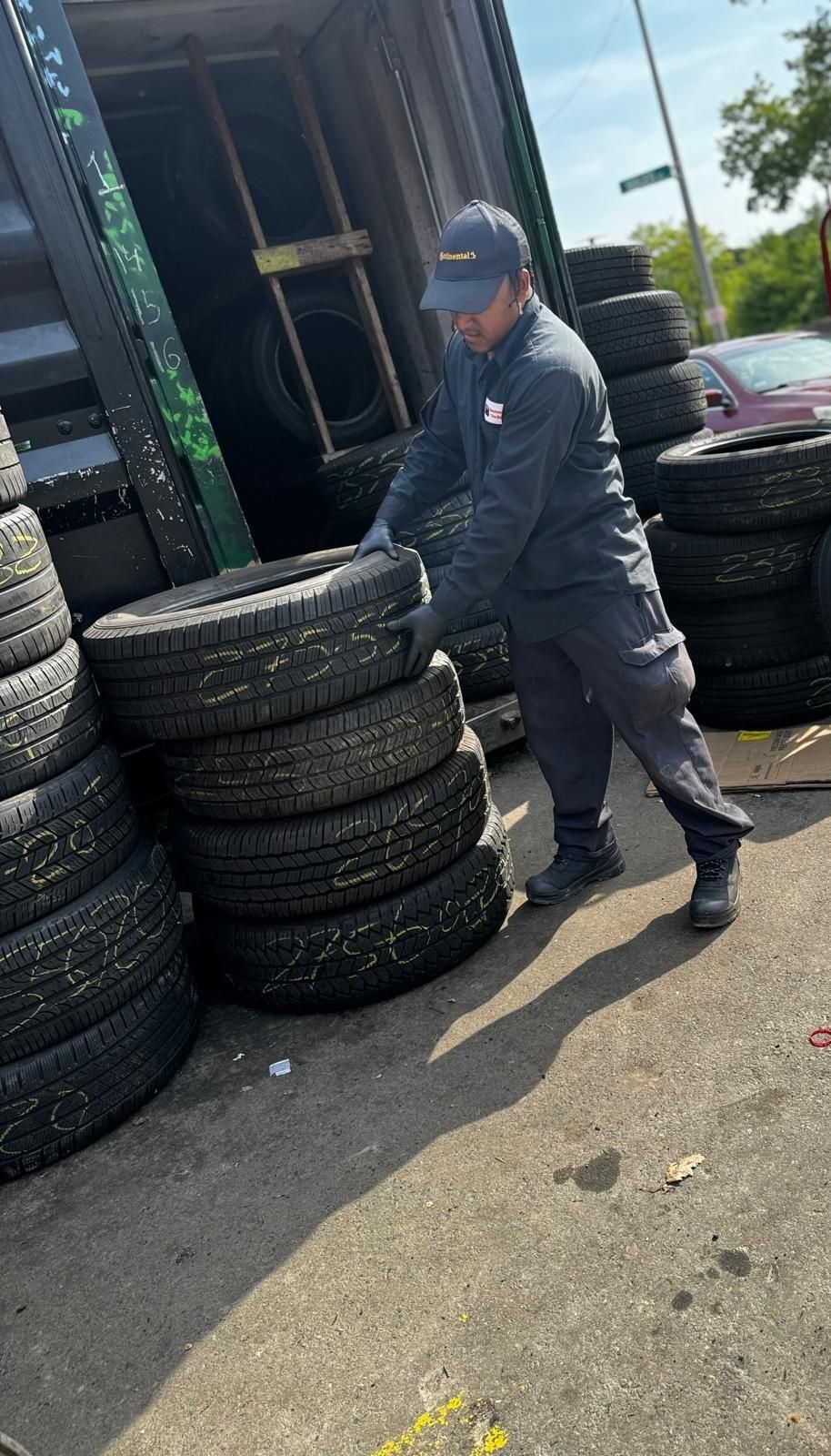 Worker stacking tires beside a red cart outside a building on a sunny day