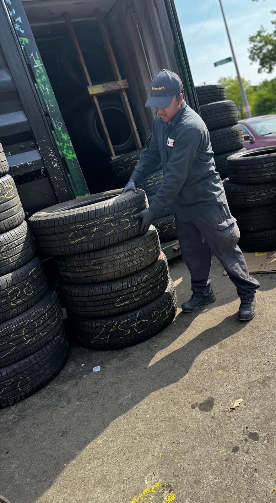 Worker stacking tires beside a red cart outside a building on a sunny day
