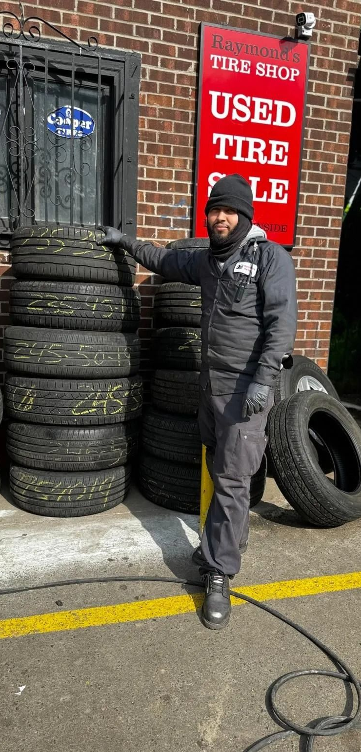 Man stands by tires stacked outside a used tire shop. Shop has a red sign, brick building.