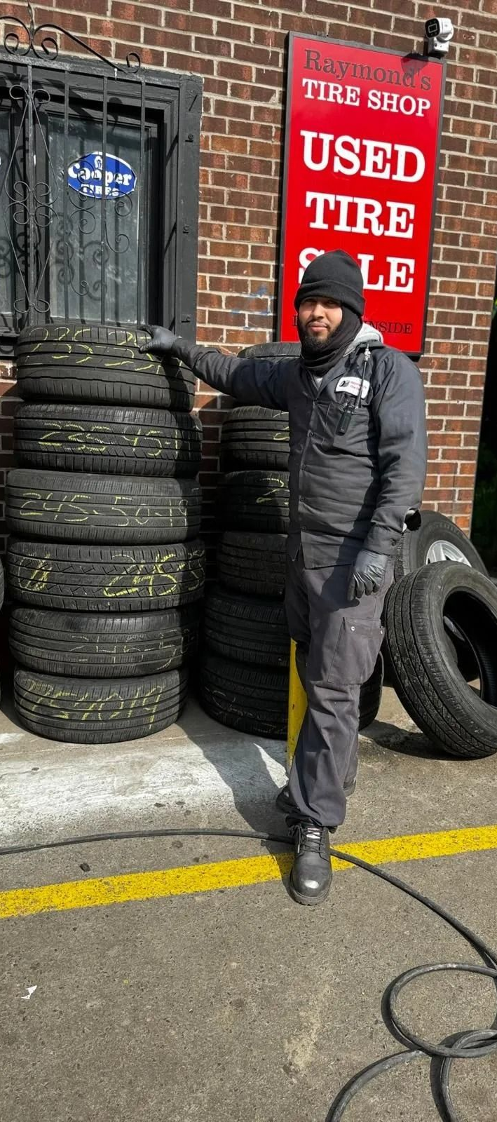Man stands by tires stacked outside a used tire shop. Shop has a red sign, brick building.