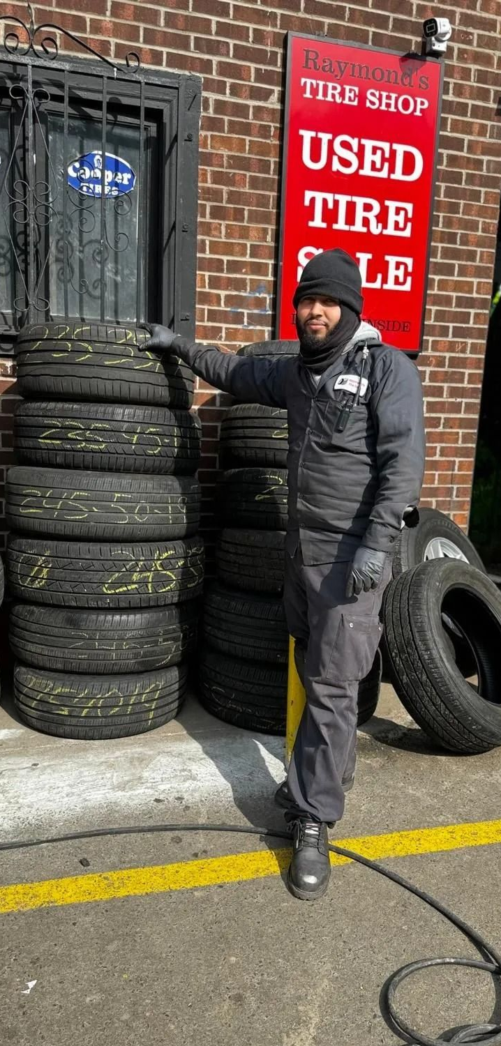 Man stands by tires stacked outside a used tire shop. Shop has a red sign, brick building.