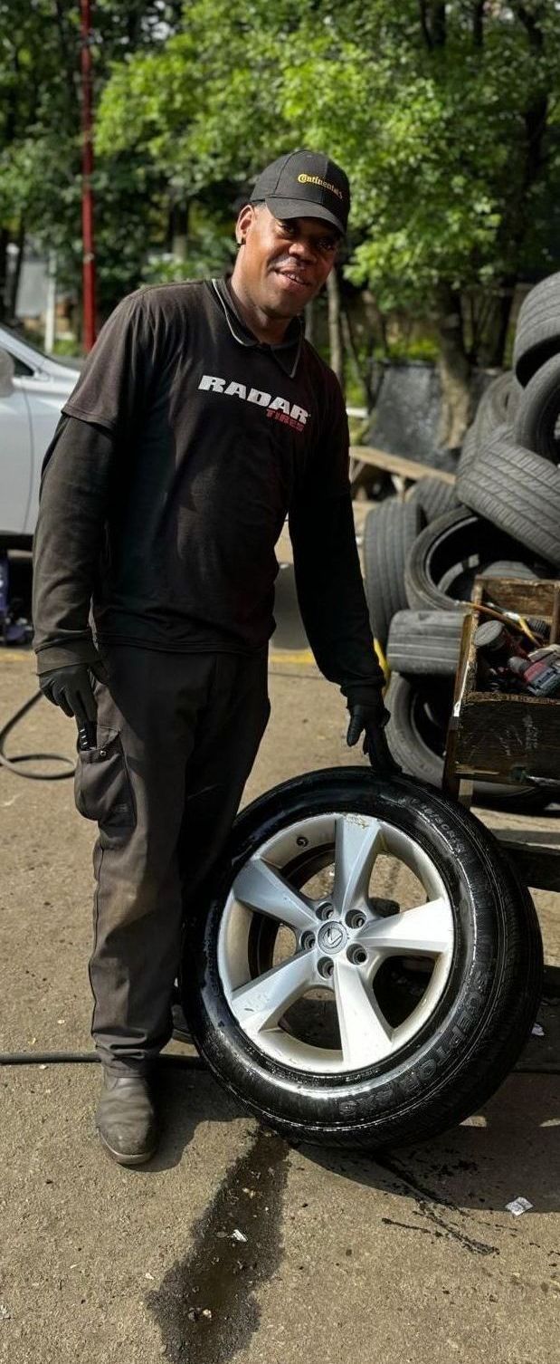 Man in work clothes holds a car tire and rim. He is outside, near a pile of tires.