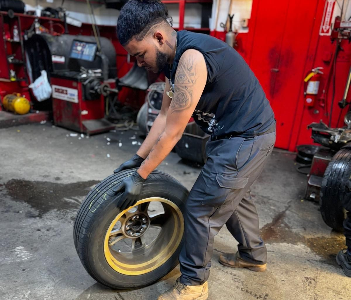 Mechanic handling a tire, inside a garage. He is wearing gloves and a black shirt.