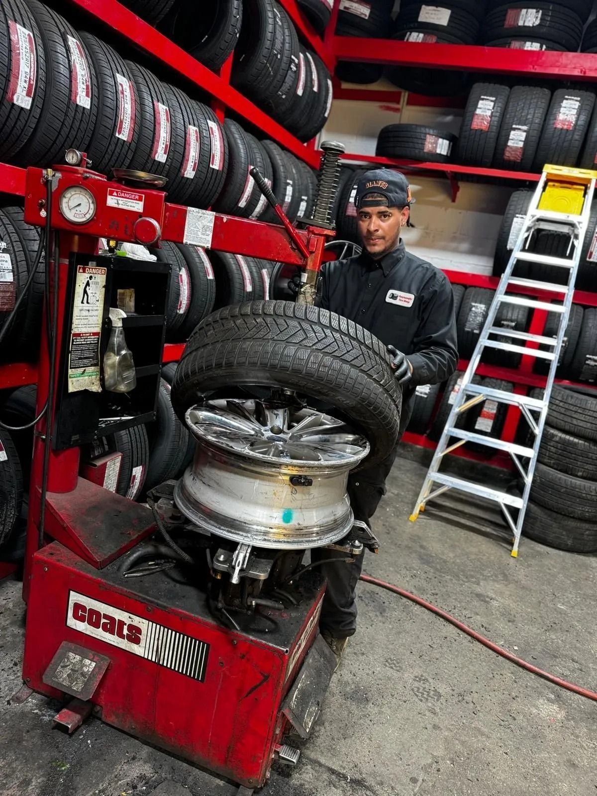 Mechanic using a tire-changing machine in a tire shop, with stacked tires and a ladder in the background