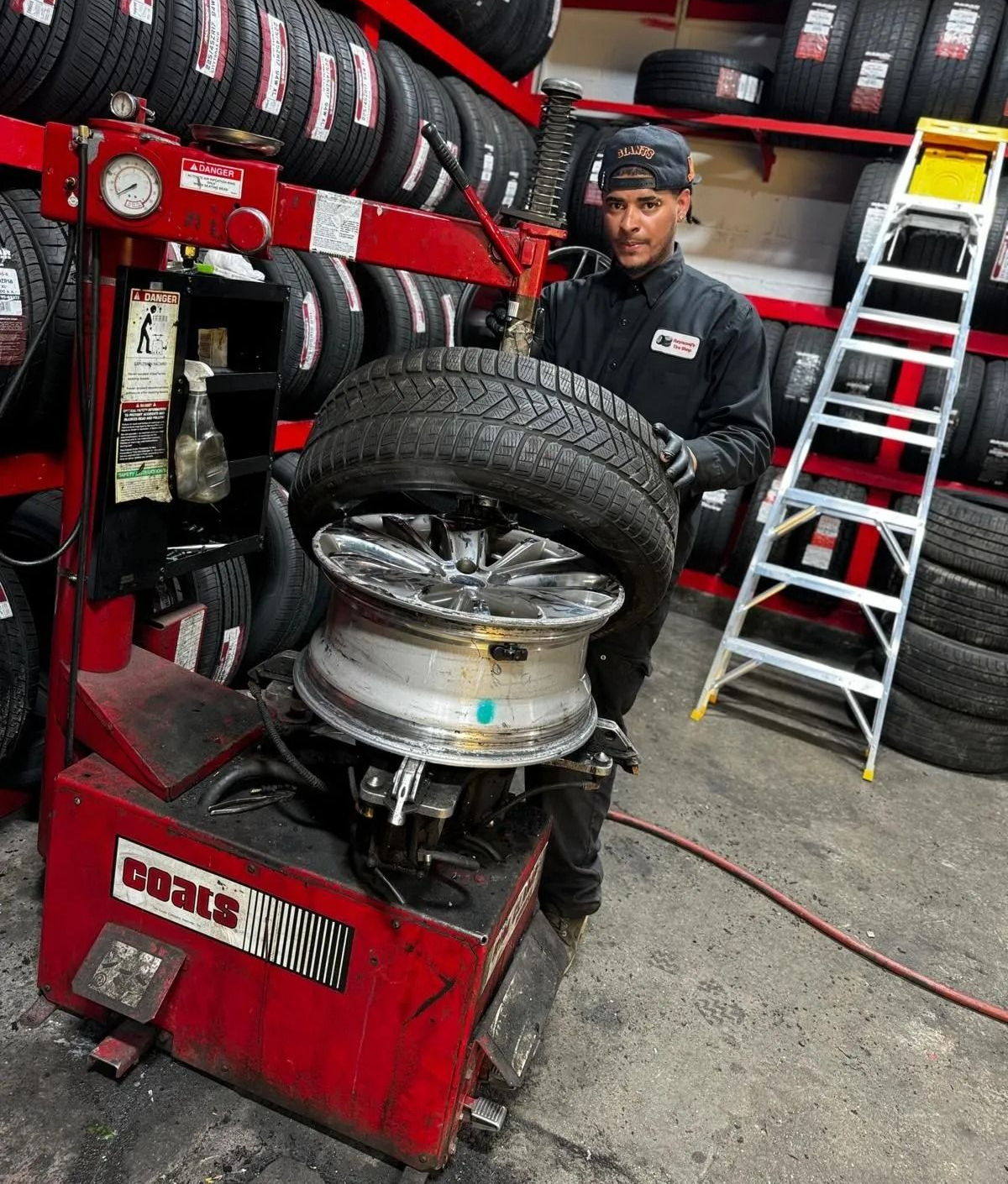 Mechanic using tire changer on a car wheel in a tire shop with stacked tires and tools nearby