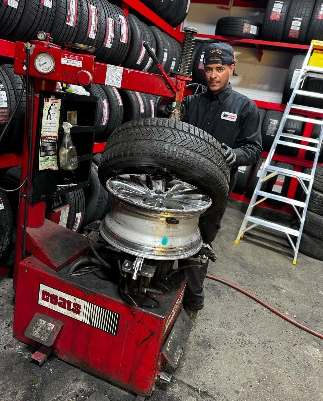Mechanic mounting a tire on a wheel in a busy auto shop, with stacked tires and tools nearby