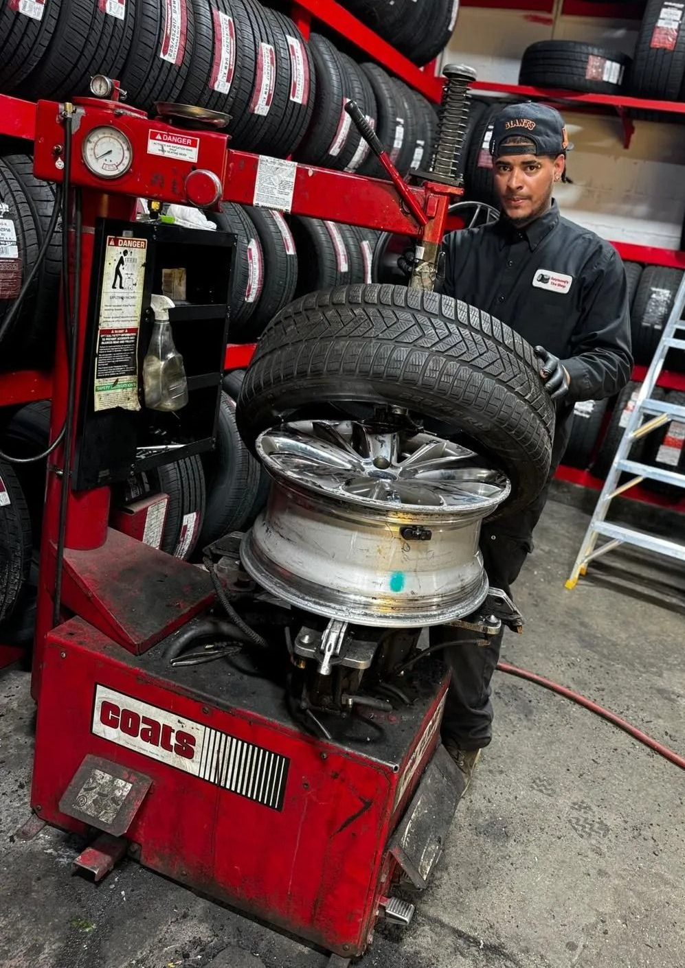 Mechanic mounting a tire on a wheel in a busy auto shop, with stacked tires and tools nearby