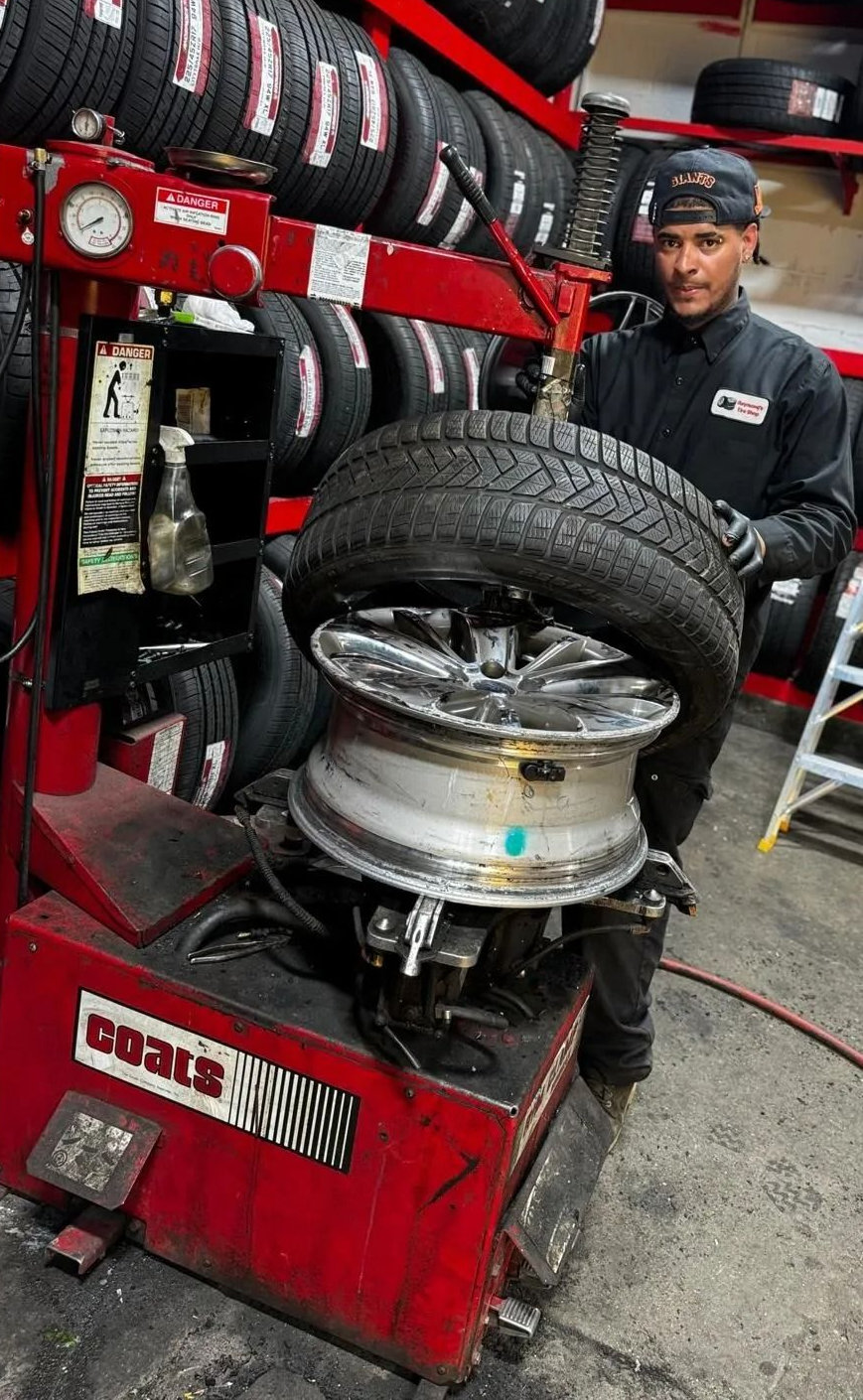 Mechanic mounting a tire on a wheel in a busy auto shop, with stacked tires and tools nearby