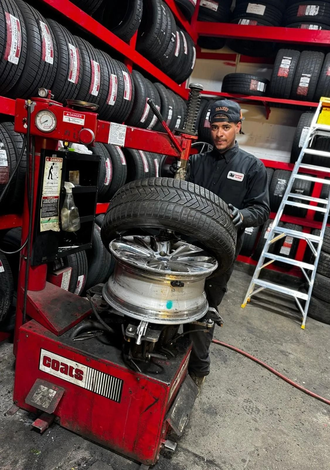 Mechanic using a tire-changing machine in a tire shop, with stacked tires and a ladder in the background