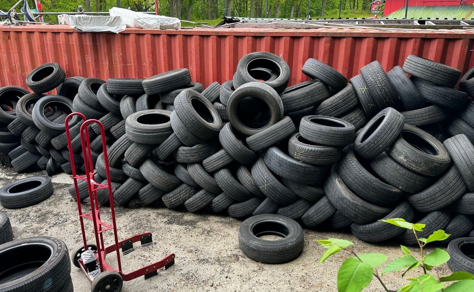 Pile of used tires stacked against a red container, with a red hand truck in front and trees behind