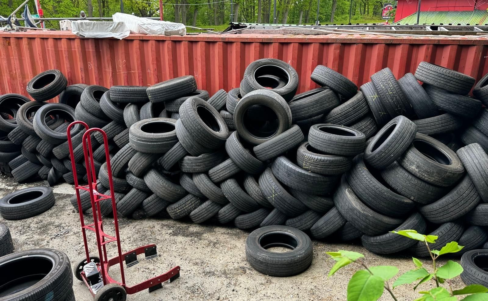 Stacked tires piled against a red shipping container outdoors, with a red dolly in front.