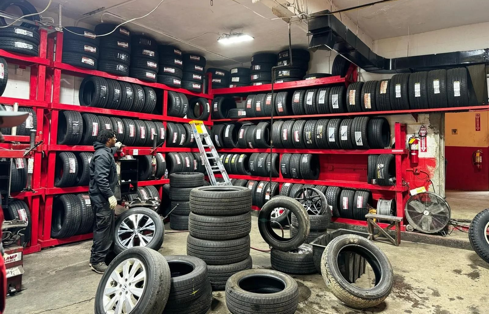 A mechanic in a tire shop surrounded by stacked tires on red shelving.
