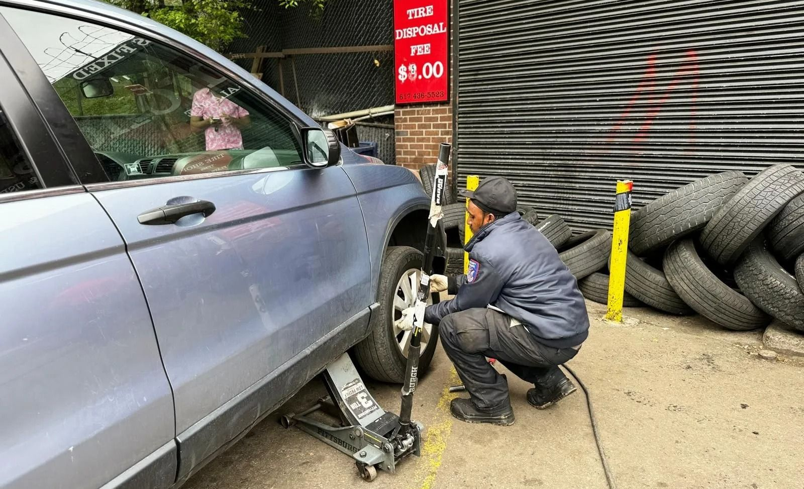 Mechanic kneels by a silver car’s front wheel, changing a tire beside stacked tires in a garage.