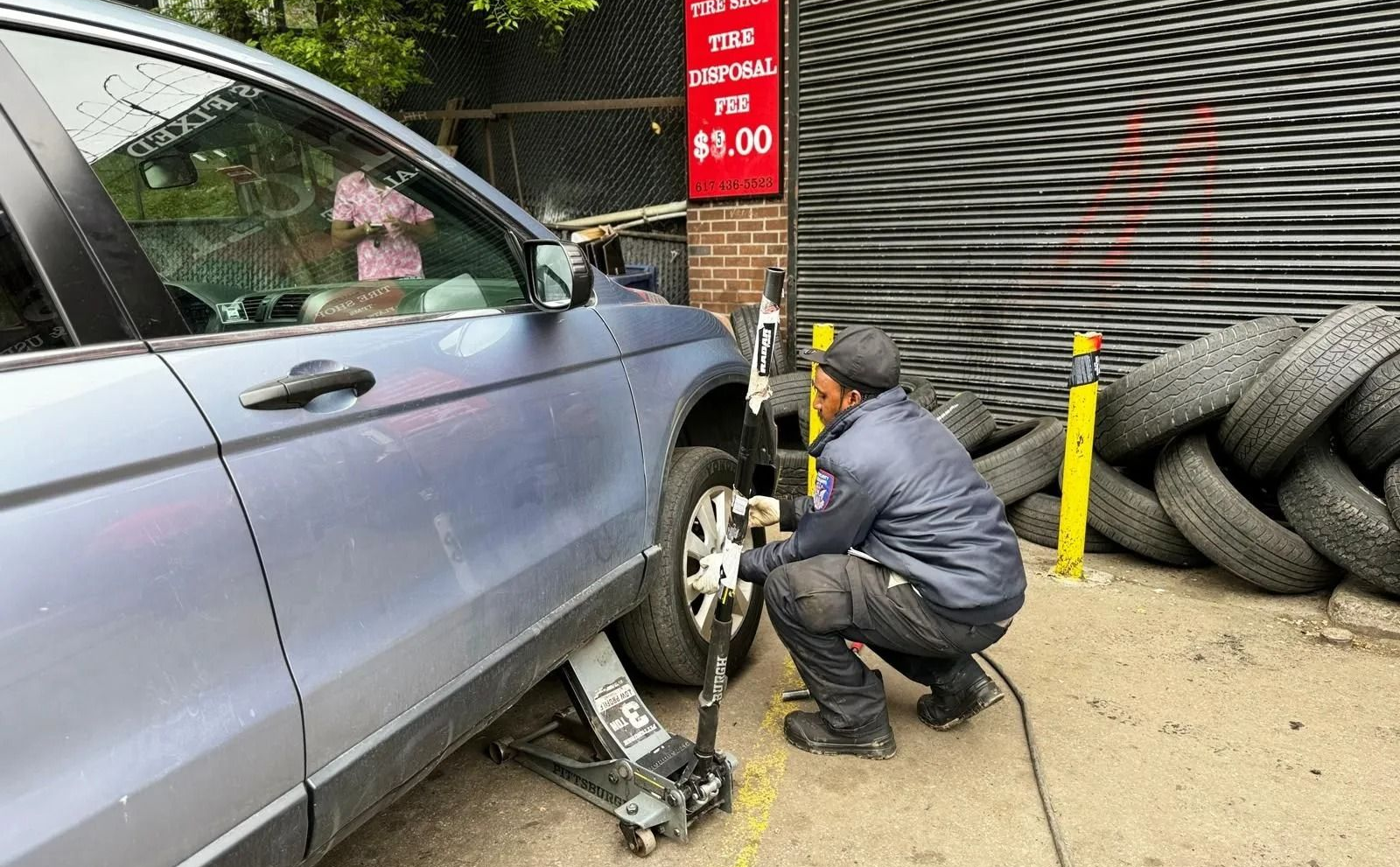 Mechanic crouches beside a blue car’s front wheel, working with a jack in an auto shop.