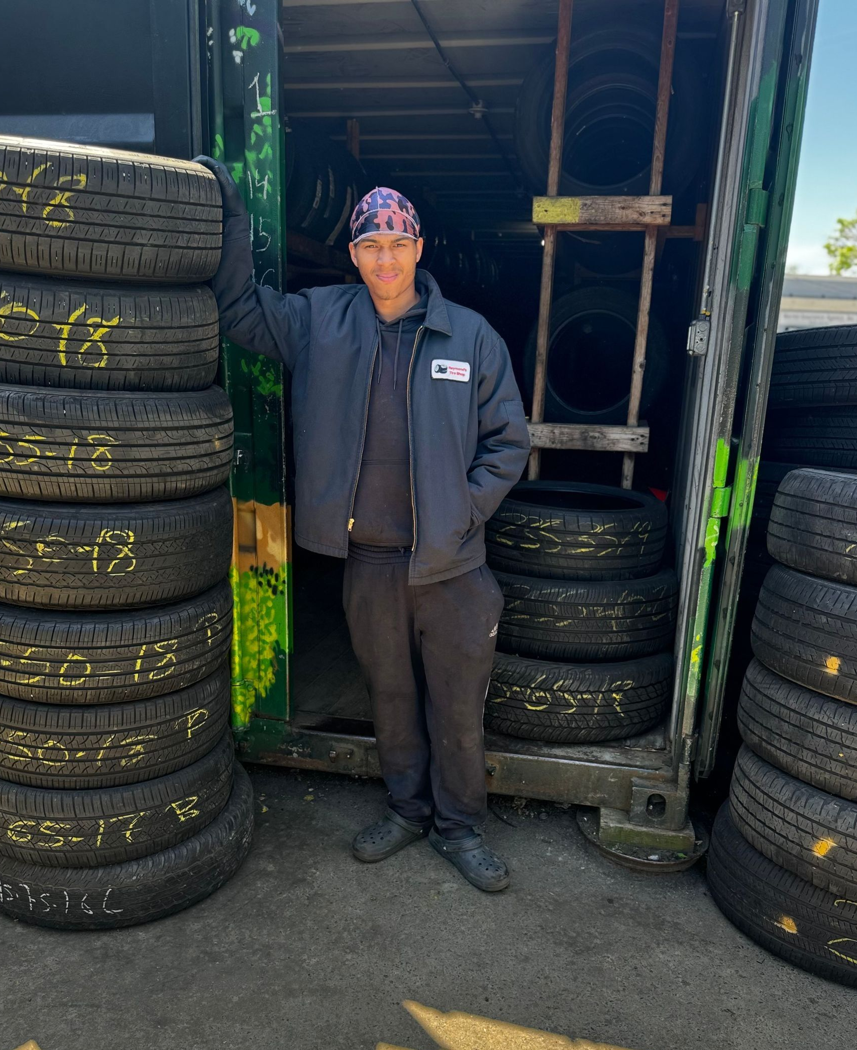 Person standing between stacks of tires in a workshop doorway, wearing a dark jacket and cap.