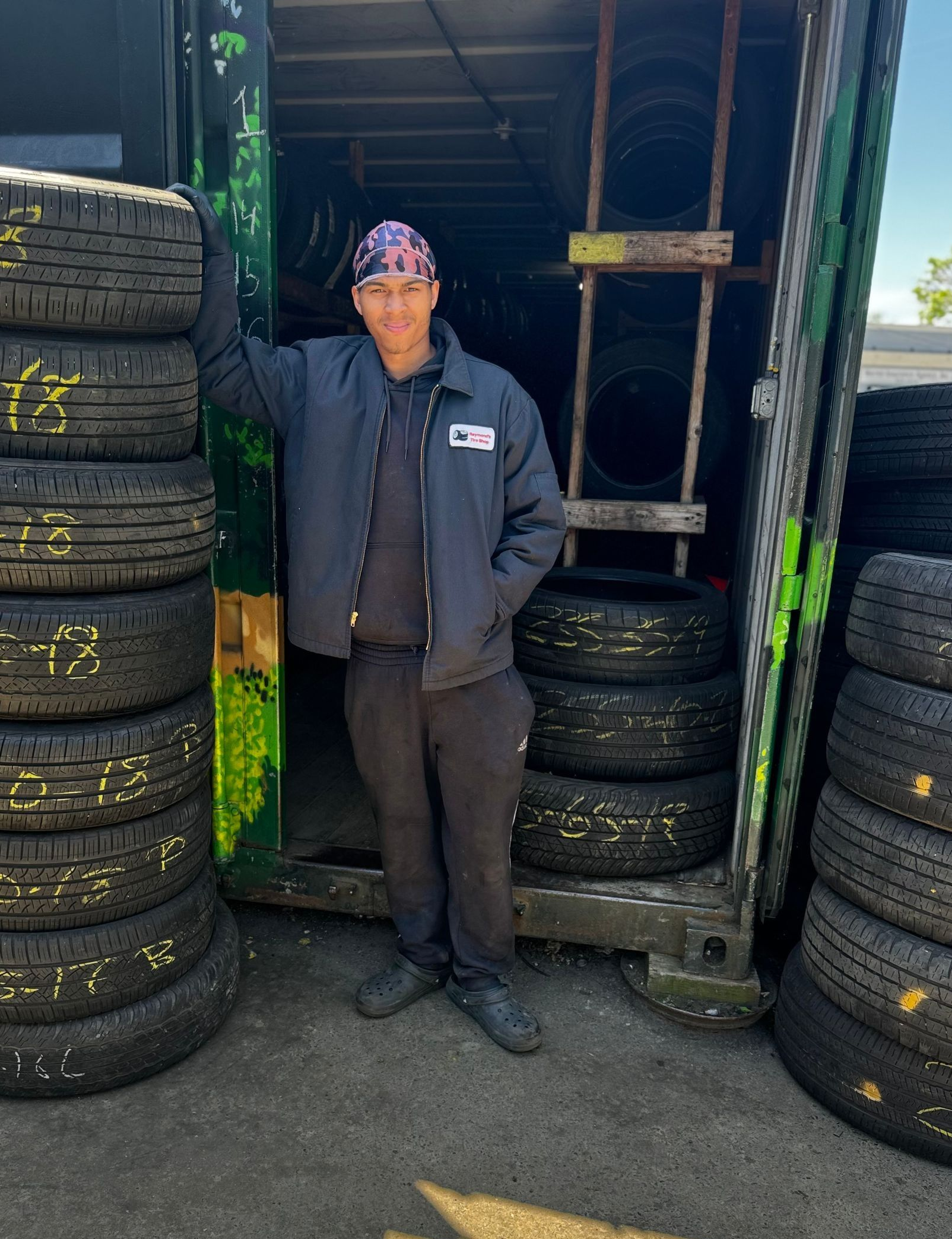 Person standing between stacks of tires in a workshop doorway, wearing a dark jacket and cap.