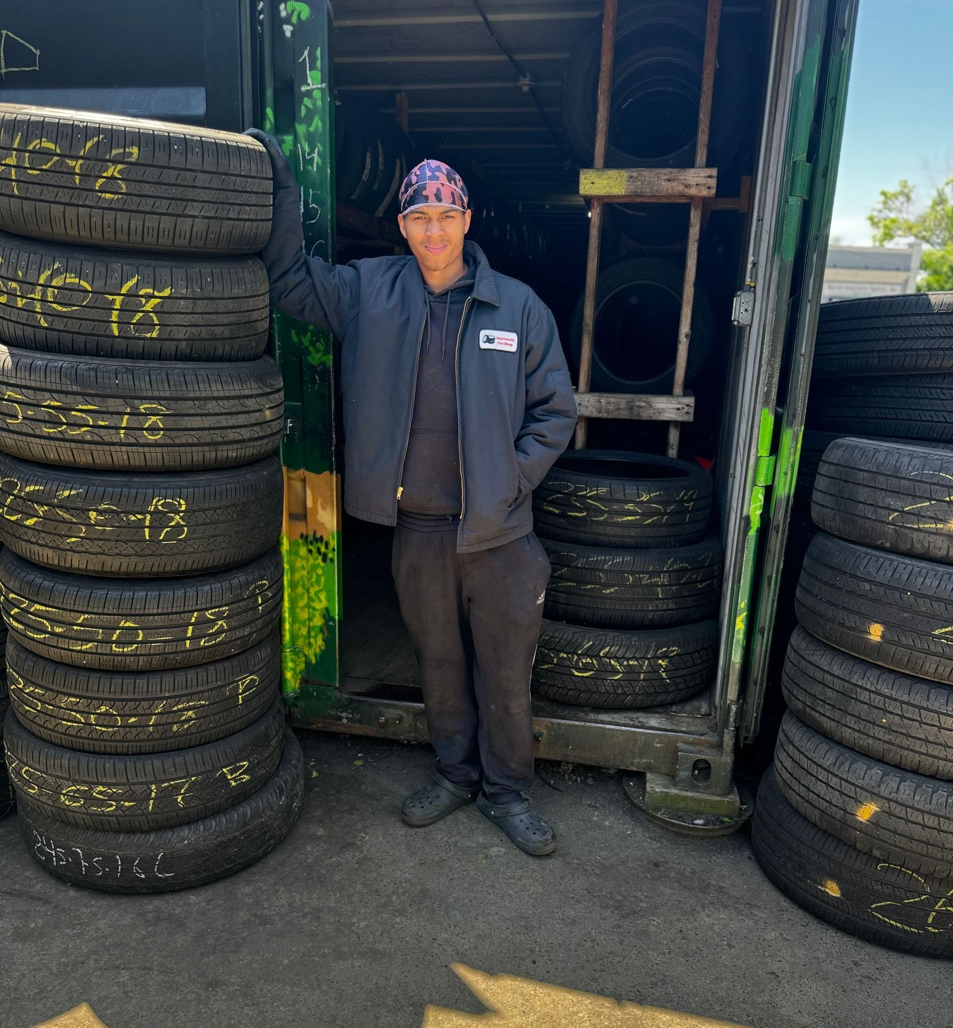 Person standing between stacks of tires in a workshop doorway, wearing a dark jacket and cap.