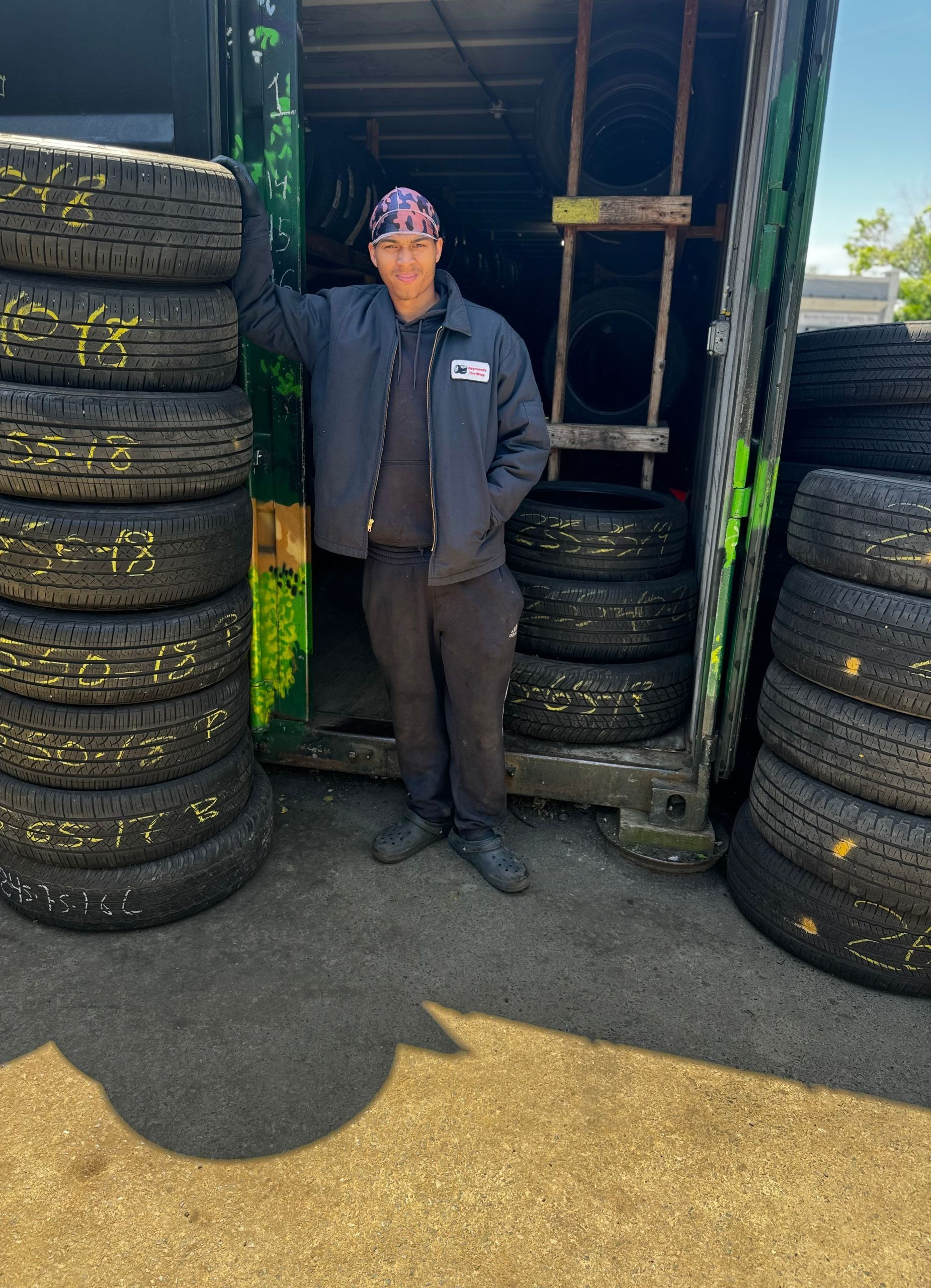 Person standing between stacks of tires outside a shop, wearing a dark jacket and cap.