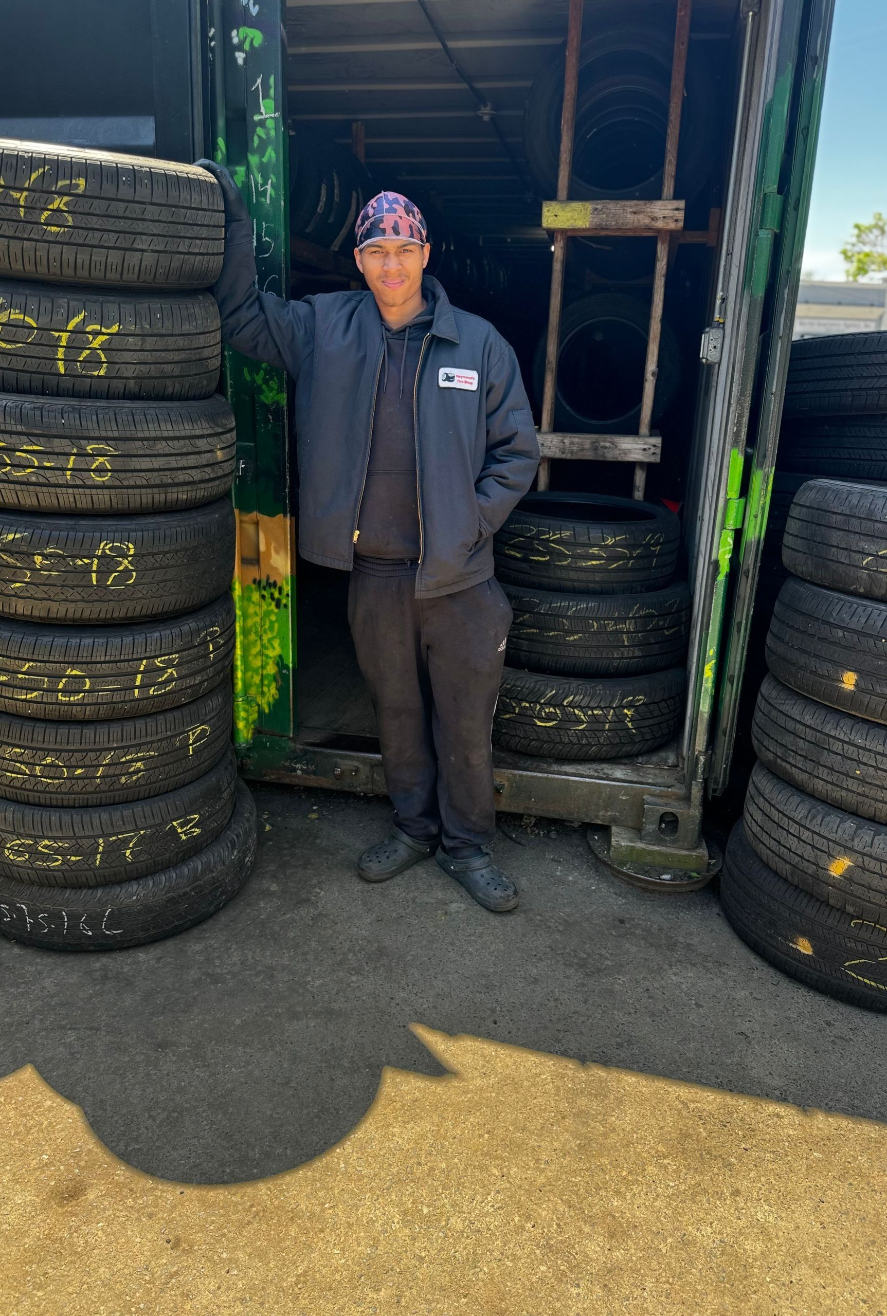 Person standing between stacks of tires outside a shop, wearing a dark jacket and cap.