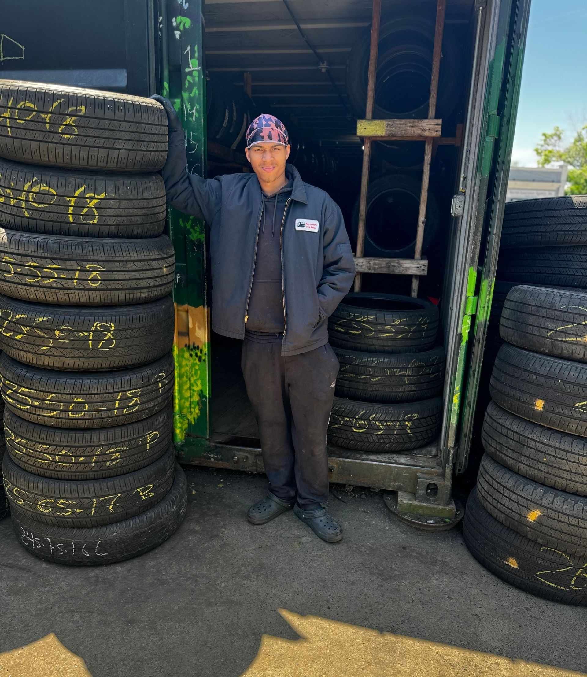 Person standing between stacks of tires outside a shop, wearing a dark jacket and cap.