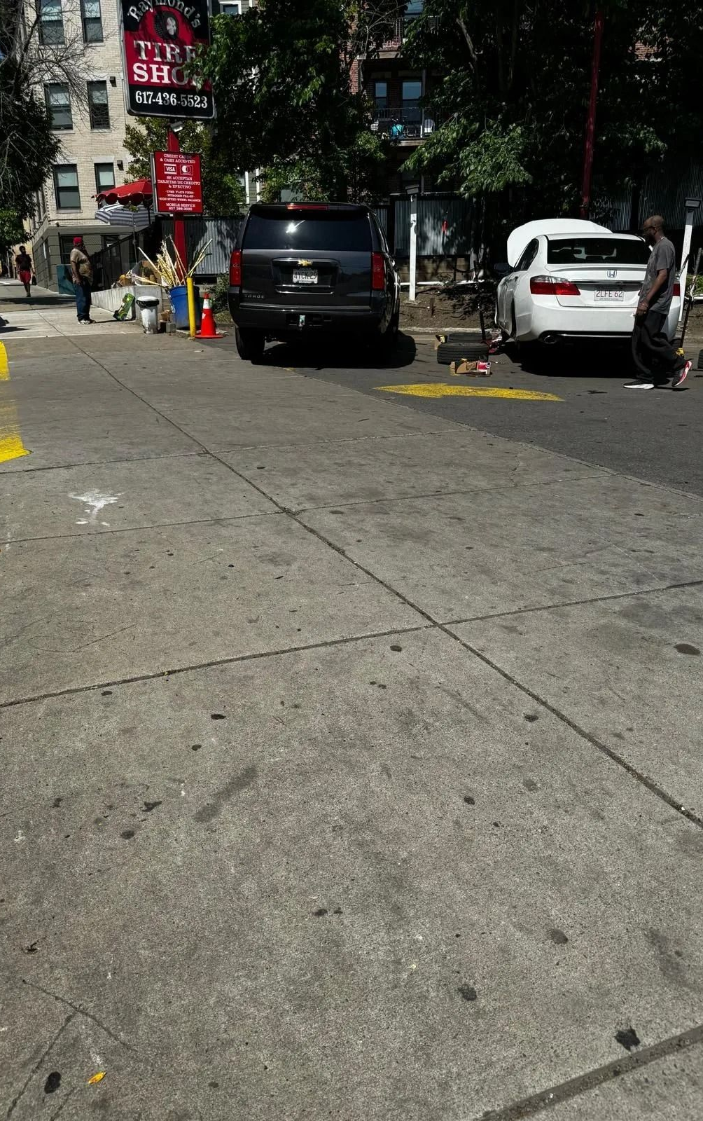 Urban street with parked black and white cars, trees, and a bright sidewalk in sunlight.