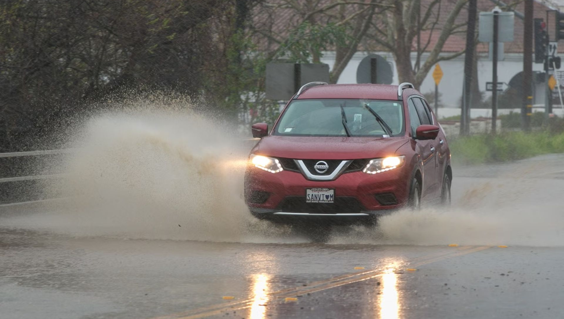 Red SUV driving through a large puddle on a wet road, spraying water.