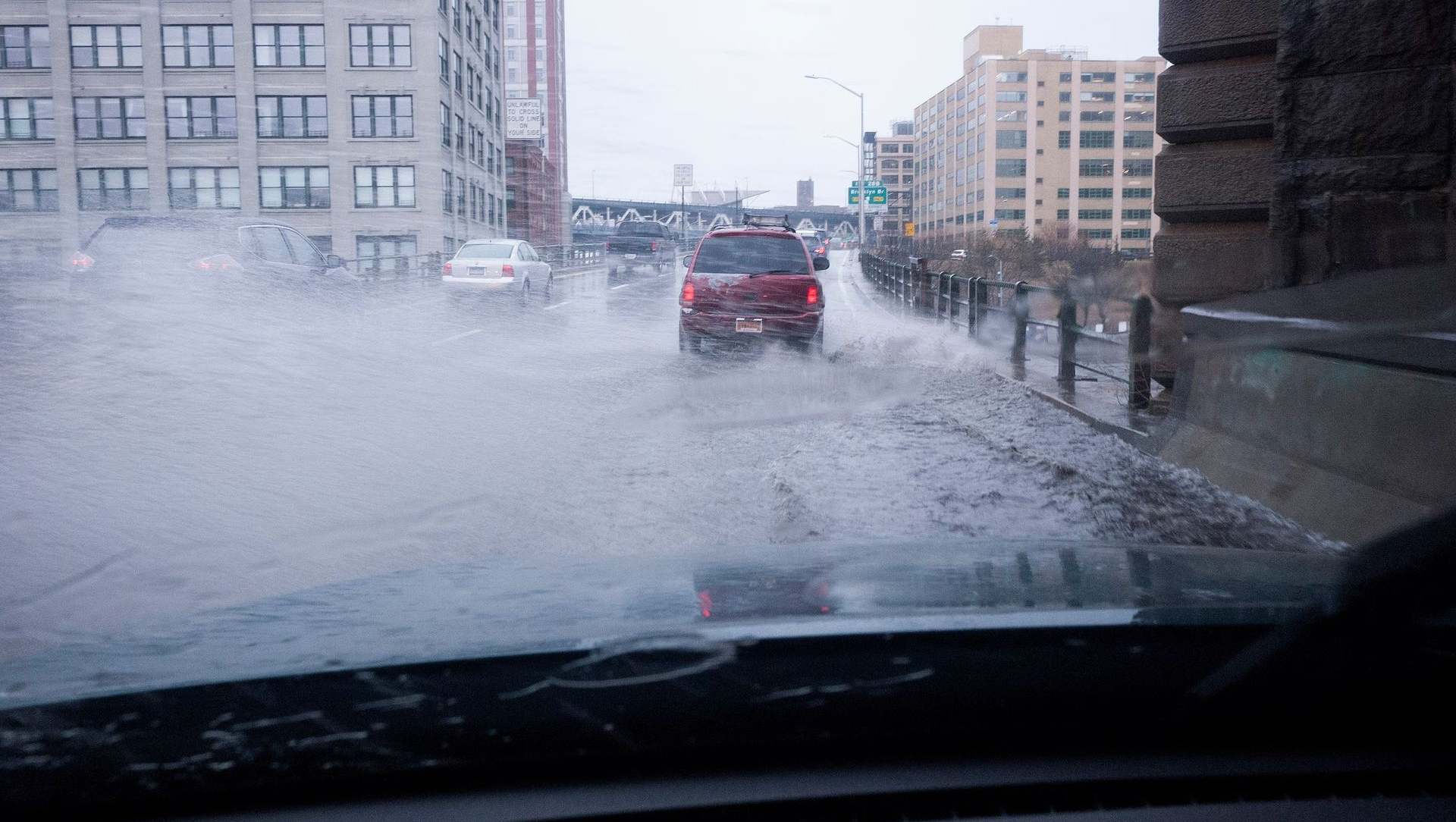 Car driving on a flooded street in a city, water splashing up. Gray sky, buildings in background.