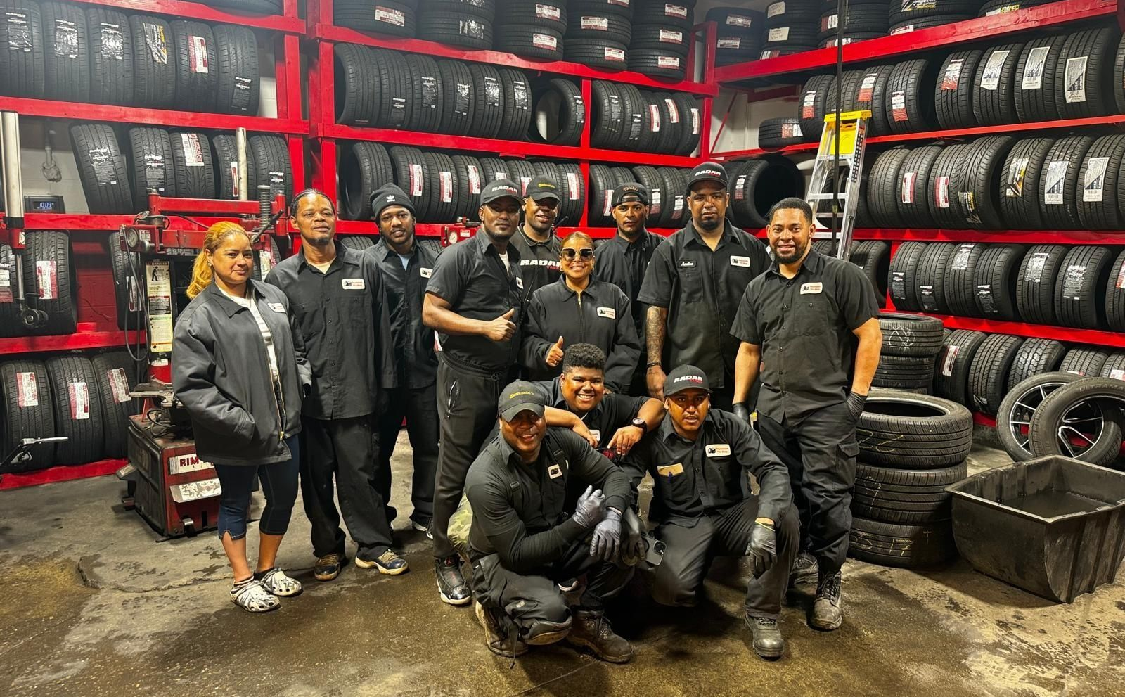 Group of people posing in a tire shop, surrounded by stacked tires and red shelving.